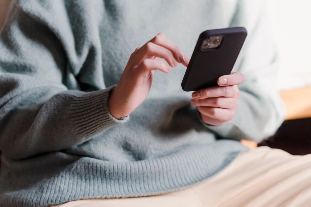 Crop faceless female in casual clothes using smartphone and touching screen while chilling at home
